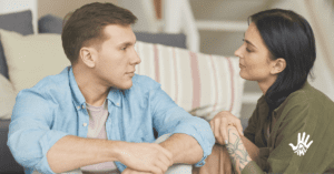 Homem loiro de cabelos curtos, vestido em camisa de botão azul clara está sentado com as mãos apoiadas nos joelhos. Ele olha para a esquerda. À sua frente, vestida em uma blusa verde musgo, está uma mulher, também de perfil, olhando-o.