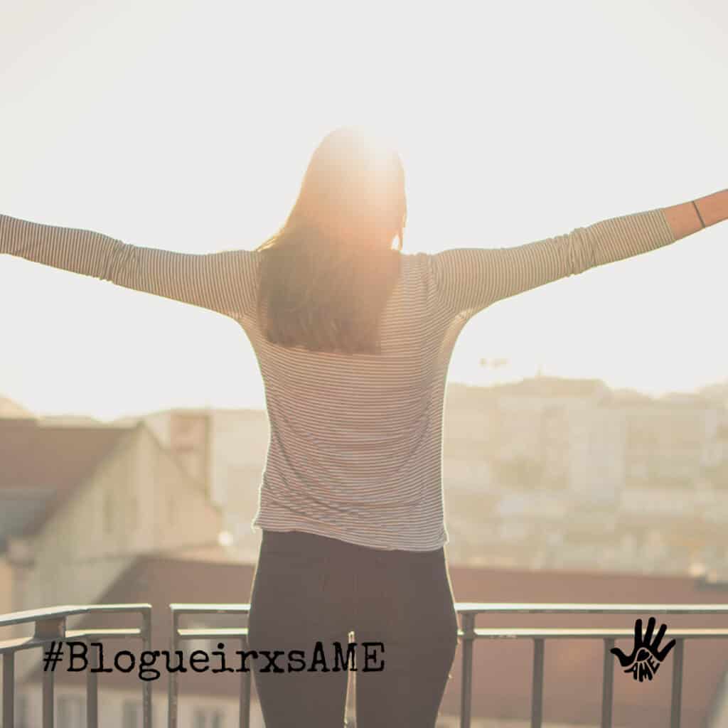 De costas, mulher de cabelos compridos está com os dois braços abertos, perpendiculares ao corpo, sua frente virada para o por do sol. No canto inferior esquerdo "#BlogueirxsAME".