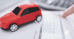 No lado esquerdo, mãos brancas em cima de um papel preenchido com tabelas. À direita, um carro vermelho miniatura em cima dos papeis.