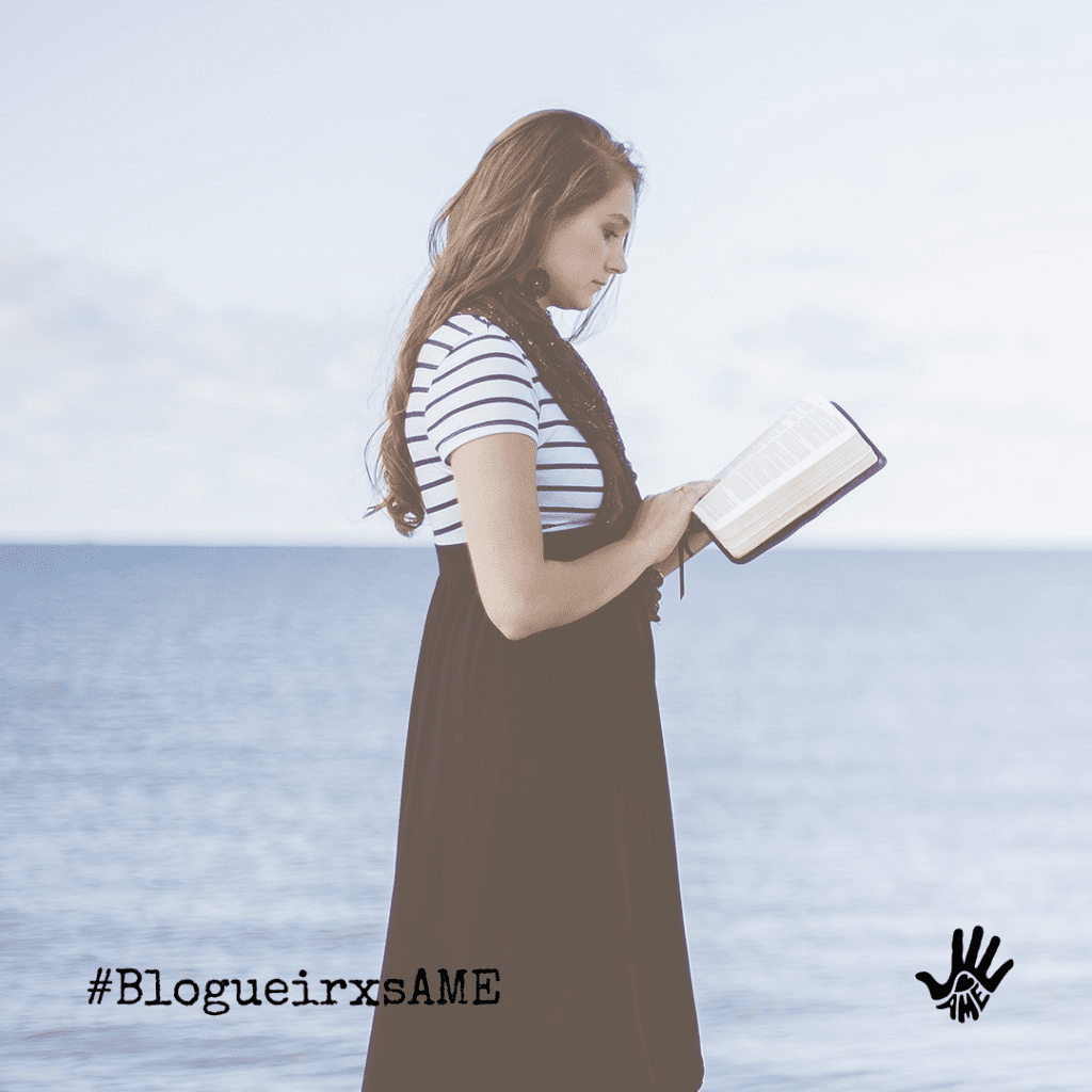 Mulher de blusa de mangas curtas listrada de branco e preto e macacão preto segura um livro nas mãos. Ela está em pé, de perfil. Ao fundo, o mar azul.