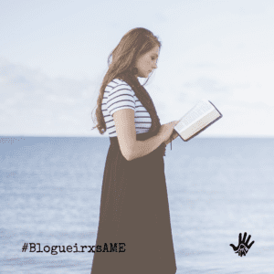 Mulher de blusa de mangas curtas listrada de branco e preto e macacão preto segura um livro nas mãos. Ela está em pé, de perfil. Ao fundo, o mar azul.