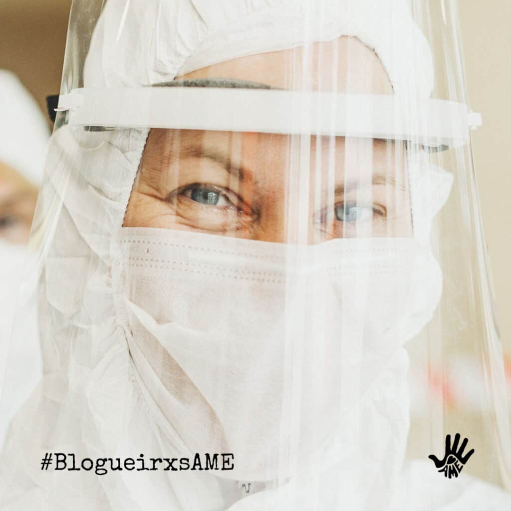 Olhos apertados fitam a câmera. Pessoa está com touca branca, máscara branca que tapa o rosto todo, menos os olhos, e face shield.