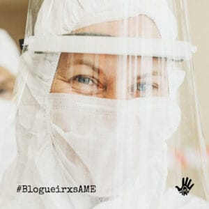 Olhos apertados fitam a câmera. Pessoa está com touca branca, máscara branca que tapa o rosto todo, menos os olhos, e face shield.