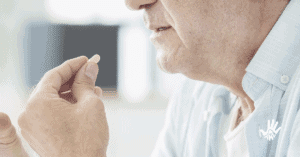 Homem de perfil, mostrando apenas a ponta do nariz, boca, queixo arredondado e levemente para frente e pescoço, segura em frente à sua boca um comprimido amarelo entre o indicador e o polegar da mão esquerda.