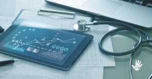 Em cima de uma mesa estão um estetoscópio preto, um tablet com a tela mostrando gráficos e papeis.