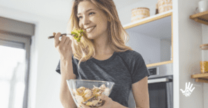 Mulher de cabelos lisos e loiros segura, com a mão esquerda, uma vasilha transparente com salada dentro. Com a mão direita, ela segura um garfo com folhas verdes levantado em frente à sua boca, que está aberta em um sorriso. Ela veste camiseta de mangas curtas cinza.