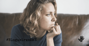 Mulher de cabelos loiros na altura do ombro, olhos azuis e pele clara, está vestida em uma camiseta cinza escura, sentada em um sofá de couro marrom. Sua mão esquerda repousa em seu queixo e ela olha para o lado esquerdo, com o rosto de perfil.