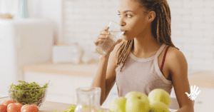 Mulher negra no canto esquerdo, de perfil, com os cabelos trançados em tranças estreitas e preso atrás da orelha, bebe água em um copo de vidro e olha para frente.