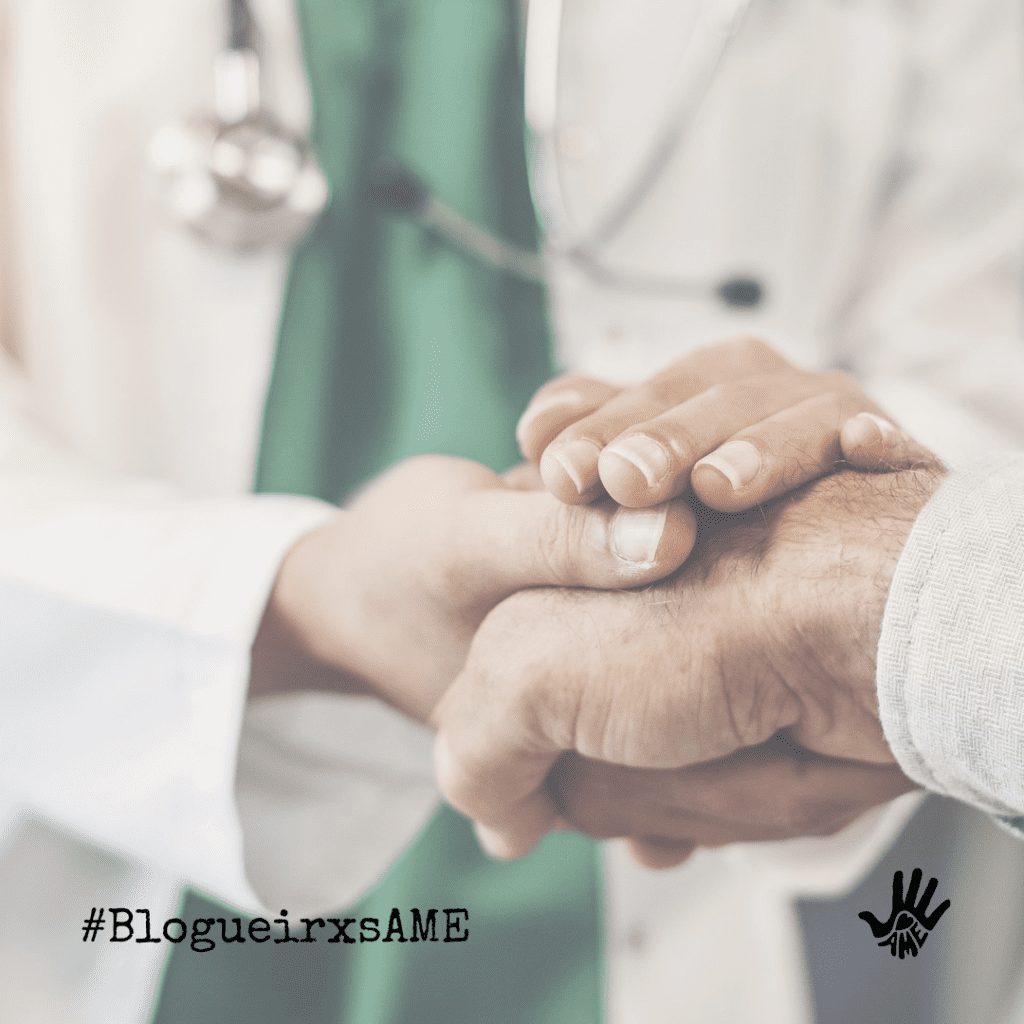 Pessoa vestida em trajes cirúgicos verdes, com um estetoscópio no pescoço e jaleco branco segura a mão de outra pessoa.