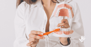 Torso de mulher vestida em uma camisa branca de botões e mangas cumpridas, em sua mão esquerda ela segura uma maquete de boca aberta com todos os dentes à mostra, com a mão direita, ela segura uma escova de dentes laranja e a coloca em contato com os dentes inferiores da maquete.