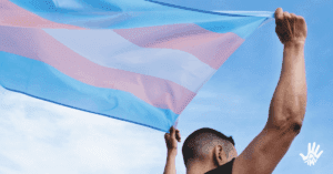 Pessoa de cabelos raspados e camisa preta, sem mangas, encontra-se de costas e com os braços levantados acima da cabeça, segurando uma bandeira listrada nas cores azul, rosa e branco.