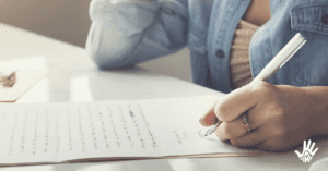 Pessoa vestida em camisa de botão jeans, aberta, mostrando o decote de uma blusa rosinha claro. Ela está sentada em uma mesa, com um braço dobrado em direção ao rosto e a outra mão segurando uma caneta que encosta em um papel sobre uma mesa branca, à sua frente.